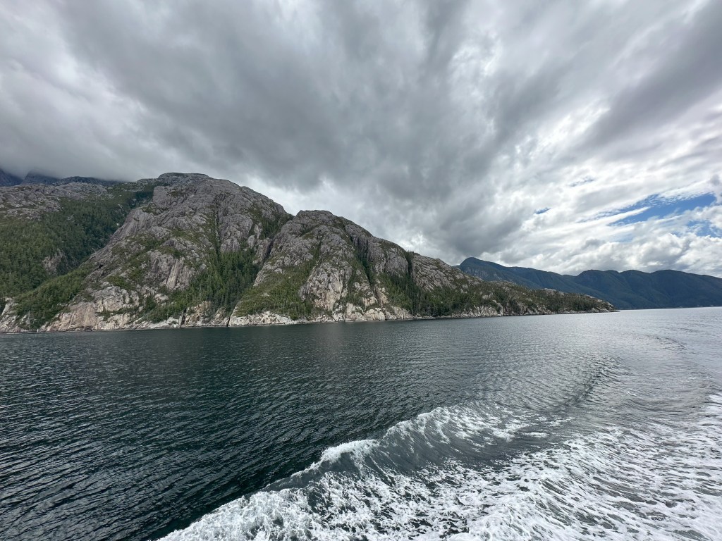 A rocky shoreline. The rocks are mostly bare with a few small trees in patches. There is a boat wake in the foreground. The sky is cloudy, with streaks of cloud appearing to radiate from the hills.