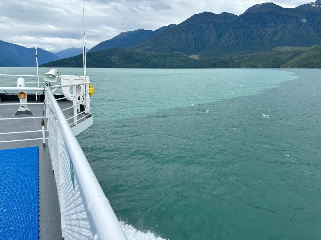 Ocean water showing a colour change where glacial flour is being deposited into the water from streams eroding the mountains. There is a clear line separating the clear ocean water from the lighter, sediment-laden water. There are mountains in the background and the bow of the ship in the left of the image.